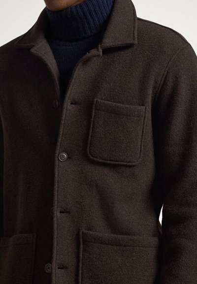 Brūns vilnas maisījuma žakete ar apkakli, priekšējiem pogām, divām uzšuvuma kabatām un teksturētu virsmu, valkāta virs tumša augsta apkakla džempera.