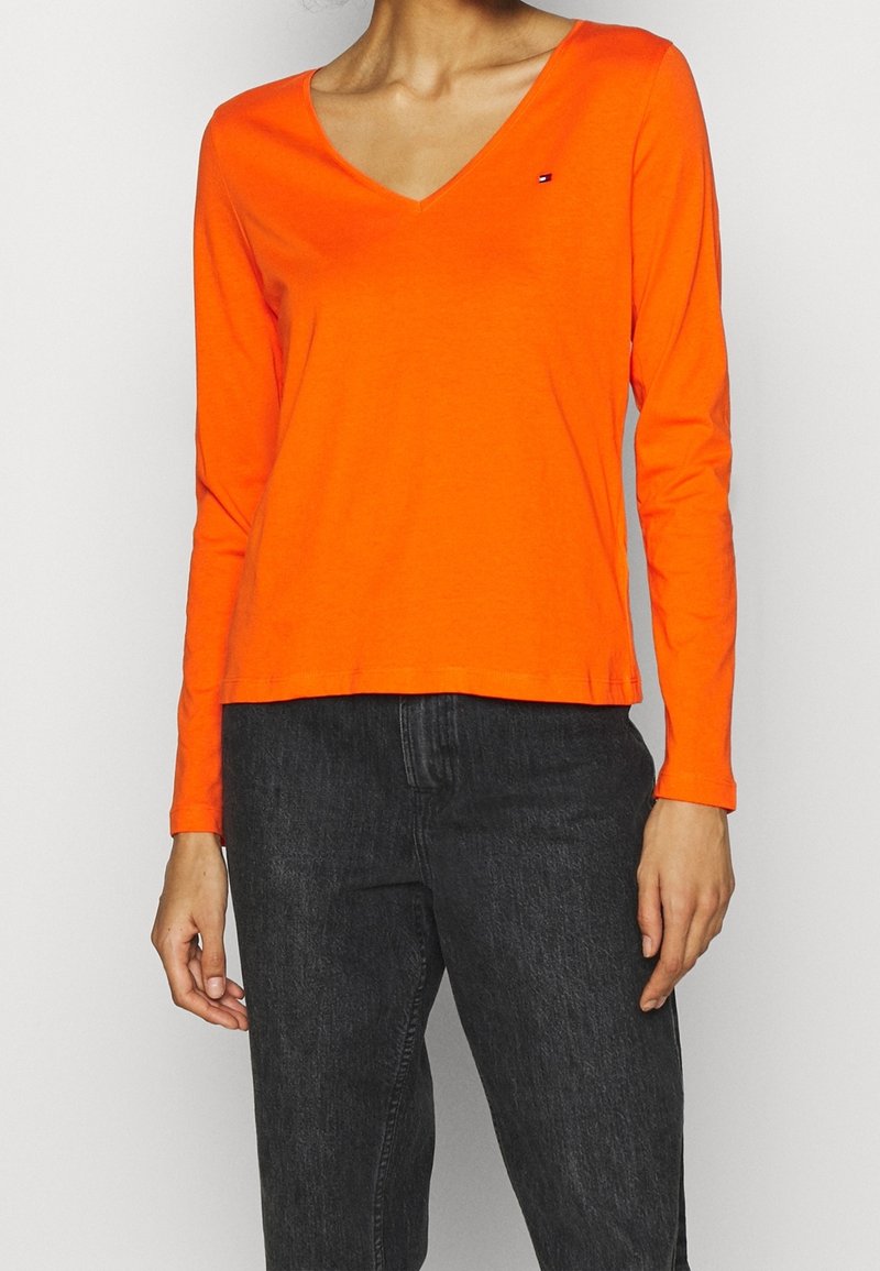 Orange langærmet V-hals skjorte lavet af bomuld, med et lille logo på brystet og en afslappet pasform, kombineret med sorte jeans.
