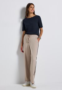 Haut à manches courtes marine associé à un pantalon de jogging beige avec des bandes latérales noires et une ceinture élastique, porté avec des baskets blanches.