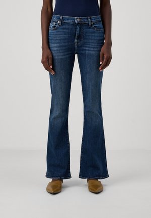 Persona con vaqueros acampanados azul oscuro, camiseta azul marino y zapatos puntiagudos color tostado, de pie frente a un fondo gris liso.