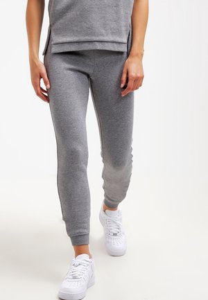 Person trägt graue Jogginghose und passendes Oberteil, läuft mit weißen Sneakers vor einem schlichten hellen Hintergrund.