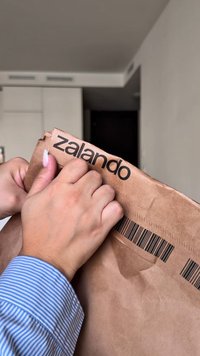 Brązowa papierowa paczka od Zalando z kodem kreskowym i logo nadrukowanym na czarno. Ręce odrywają otwarcie paczki.
