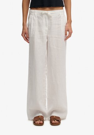 Mujer vistiendo pantalones holgados de lino blanco con bolsillos delanteros, cinturón en la cintura, top negro cropped y sandalias marrones sin talón.