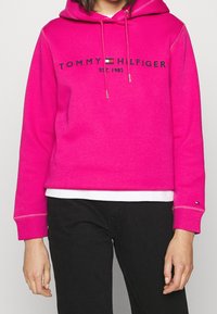 Růžová mikina s kapucí, šňůrkami a vyšitým logem "TOMMY HILFIGER". Má žebrované manžety a vrstvený bílý lem. Materiál z bavlněné směsi.