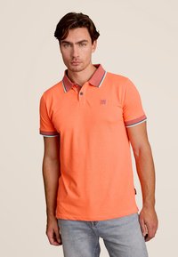 Oranje polo shirt met een gestructureerde kraag in contrasterende rode en blauwe afwerking, voorzien van een klein logo-embleem op de borst. Gecombineerd met lichte jeans.