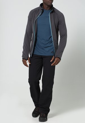 Uomo che indossa una giacca grigio scuro con zip sopra una maglietta blu, pantaloni neri e scarpe da trekking nere, in piedi davanti a uno sfondo uniforme.