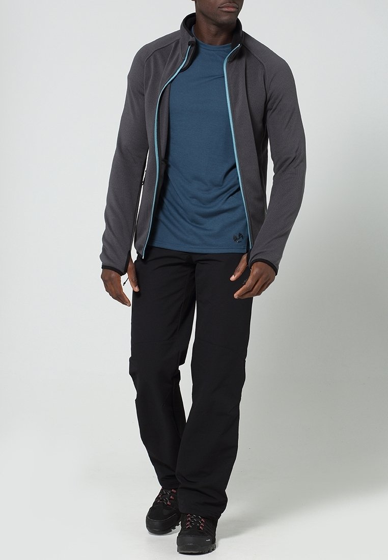 Homme portant une veste zippée gris foncé sur un t-shirt bleu, un pantalon noir et des chaussures de randonnée noires, debout devant un fond uni.