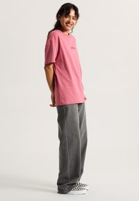 Camiseta de algodón rosa con mangas cortas, que presenta aberturas laterales y un logo en texto. Combinada con pantalones vaqueros grises holgados y zapatillas con un patrón en blanco y negro.