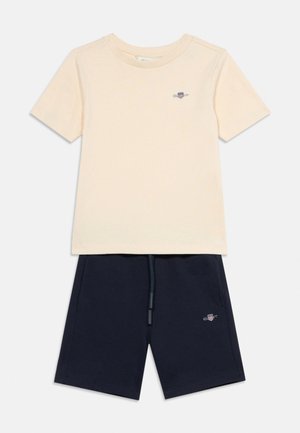 Beige kortærmet T-shirt med lille logo på brystet, kombineret med navyblå shorts med snøre og matchende logo på benet.