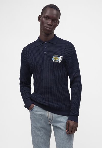 Homme portant un polo à manches longues texturé bleu marine avec plusieurs logos Moschino sur la poitrine, associé à un jean bleu clair.