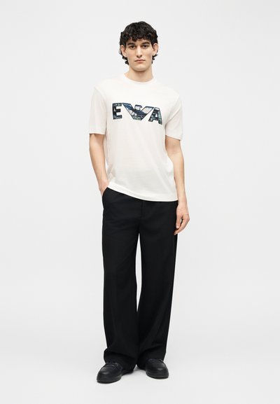 Νεαρός με σγουρά σκούρα μαλλιά φοράει λευκό πουκάμισο με κοντά μανίκια και στάμπα «EVA», μαύρο παντελόνι με φαρδιά μπατζάκια και μαύρα παπούτσια, στέκεται με το ένα χέρι στην τσέπη.