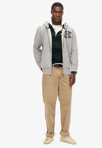 Felpa grigia con zip e tasche frontali, indossata sopra un polo a righe scure, abbinata a pantaloni beige e sneakers chiare. Accentata con un orologio.