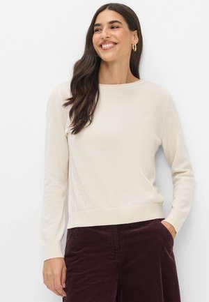Femme aux longs cheveux foncés souriante, portant un pull crème et un pantalon en velours marron foncé, debout devant un fond blanc.