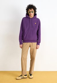 Lilla hættebluse med frontlomme og logo, beige bukser og gule sneakers med sorte striber. Modellen bærer solbriller.