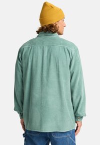 Green corduroy shirt with back pleat, long sleeves, and buttoned cuffs, paired with a yellow knit beanie. Textured surface with vertical stripes.