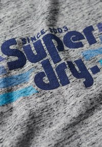 Gray fabric t-shirt featuring "Superdry" printed in bold blue, with lighter blue waves and the text "Since 2003" above the logo.