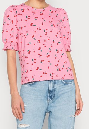 Kobieta w różowej koszulce z krótkim rękawem w czerwone wisienki, zestawionej z jasnoniebieskimi poszarpanymi dżinsami.