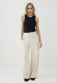 Pantaloni larghi color beige chiaro con vita regolabile, dotati di tasche laterali e texture liscia. Abbinati a un top aderente nero.