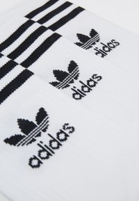 Hvite sokker med tre svarte horisontale striper på toppen. Har en svart brodert Adidas-logo med kløverdesign. Strukturert stoff.