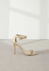Guld metallic højhælede sandaler med ankelrem, åben tå og minimalistisk design. Glat tekstur og slank stiletto hæl.