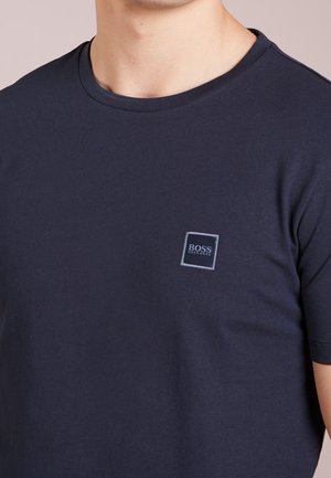 Mężczyzna w granatowej koszulce Hugo Boss z okrągłym dekoltem i małą naszywką z logo na lewej piersi.