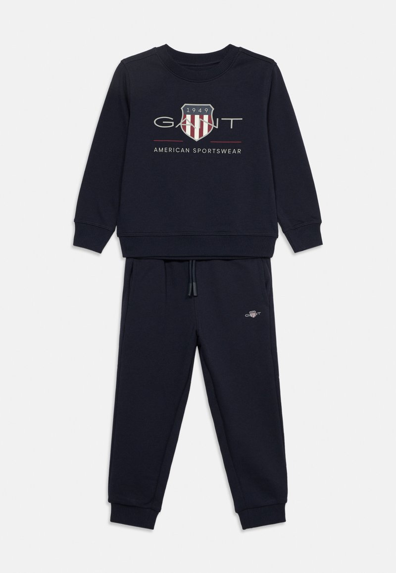 Σετ μπλε ναυτικού χρώματος φούτερ και παντελόνι φόρμας με το λογότυπο GANT και έμβλημα ασπίδας σε κόκκινο, λευκό και γκρι στη μπροστινή πλευρά του φούτερ, με ελαστικά μανσέτα και μέση.