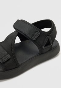 Svart sandal med stoffstropper og borrelås, med en flat gummisåle og teksturerte stoffer for komfort og passform.