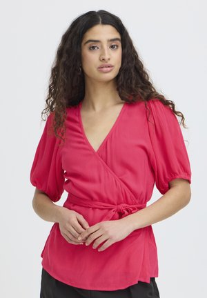 Vrouw met lang, krullend haar die een felroze wikkelblouse met opgeblazen mouwen draagt, staand tegen een effen lichte achtergrond.