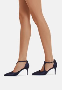 Tacchi alti in satin blu navy con punta affusolata, cinturino alla caviglia e design ritagliato. Presentano un sottile tacco stiletto e una texture liscia.