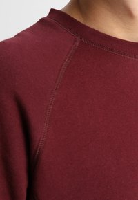 Vínově červená mikina s kulatým výstřihem, zdobená prohnutými švy a měkkou texturou. Detailní záběr na výstřih a šev na rameni.