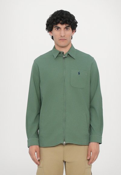 Maglietta verde con zip, colletto a camicia, taschino frontale e zip visibile, realizzata in un tessuto liscio con una vestibilità rilassata.