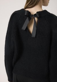 Pulover negru din tricot, cu un finisaj texturat. Prezintă o deschidere în spate legată cu o panglică din satin, decolteu rotund și umeri căzuți.