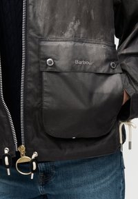 Μαύρο αδιάβροχο μπουφάν με ασημένιο φερμουάρ και κουμπιά κούμπωσης, που διαθέτει μπροστινή τσέπη με κεντημένο το λογότυπο "Barbour" και απαλή υφή.