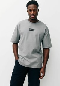 Szürke, rövid ujjú póló sima anyagból, szabályos téglalap alakú fekete logócímkével a mellkas magasságában. Sötét nadrággal párosítva.