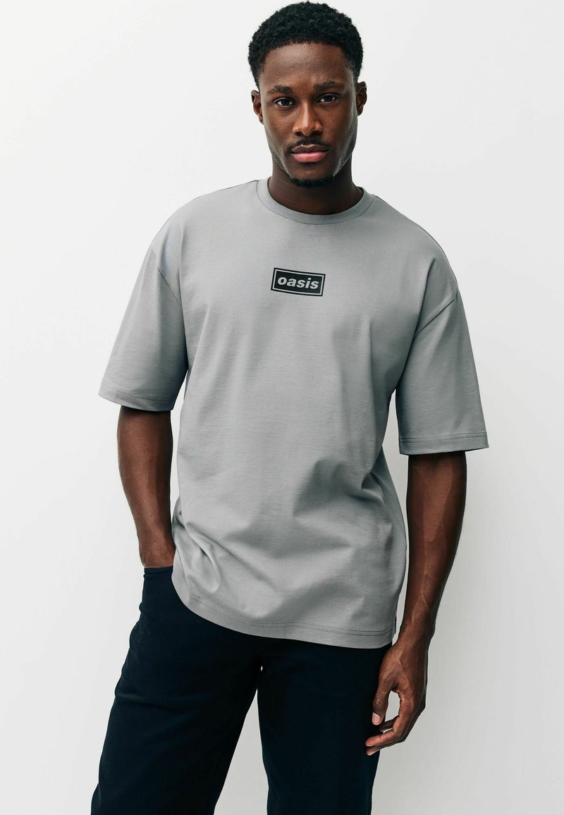 Szürke, rövid ujjú póló sima anyagból, szabályos téglalap alakú fekete logócímkével a mellkas magasságában. Sötét nadrággal párosítva.