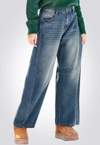 Jeans in denim blu a gamba larga con una finitura sbiadita, caratterizzati da tasche frontali e dettagli di cucitura sottili, abbinati a stivali color nocciola.