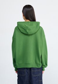 Una persona con lunghi capelli scuri indossa un grande hoodie verde, vista da dietro, su uno sfondo chiaro e semplice.