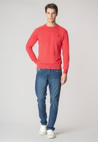 Felpa rossa lavorata a maglia con polsini e orlo a coste, abbinata a jeans in denim blu. Le scarpe sono sneakers bianche. Il modello posa con un'espressione neutra.