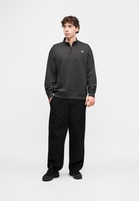 Μαύρο φλις φούτερ με φερμουάρ μέχρι τη μέση, μακριά μανίκια και λογότυπο, σε συνδυασμό με φαρδιά μαύρη φόρμα τύπου cargo και μαύρα αθλητικά παπούτσια. Απλό λευκό φόντο.