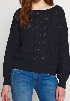 Schwarzer gestrickter Pullover mit einem lockeren Boot-Ausschnitt-Design und strukturiertem Muster, das geometrische Ausschnitte aufweist. Rippbündchen und Saumdetails.