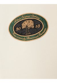 Crèmewitte stof met een geborduurd ovaal embleem met een boom, gouden tekst en groene rand. De tekst bevat "2013" en "Internationally Renowned Garments".