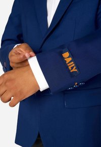 Jūras flotes zila žakete ar baltu aproci un oranžu "DAILY" etiķeti uz piedurknes. Iezīmēta ar pieguļošu piegriezumu un gludu auduma tekstūru.