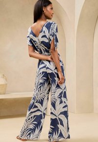 Tuta navy con motivo a foglia bianca, maniche corte e design a V sulla schiena. Presenta gambe ampie e vita arricciata, realizzata in tessuto leggero.