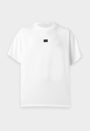 Mand iført en sort bucket-hat, hvid kortærmet T-shirt over en sort langærmet skjorte, sorte bukser og sorte sko, stående med den ene hånd i lommen.