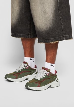 Person iført hvide Puma-sokker, grønne og bordeauxfarvede sneakers med tykke hvide såler og falmede sorte knælange denimshorts.