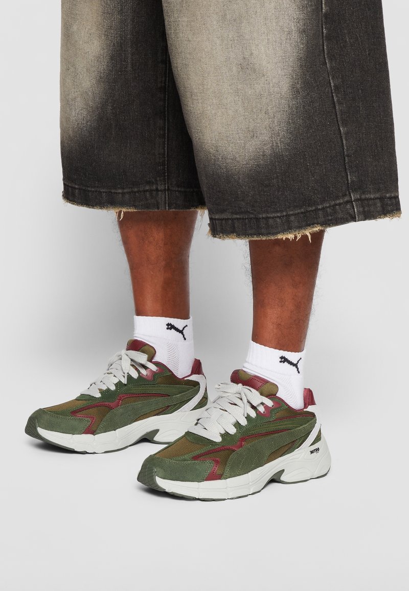 Personne portant des chaussettes blanches Puma, des baskets vertes et bordeaux avec des semelles blanches épaisses, et un short en jean noir délavé arrivant aux genoux.