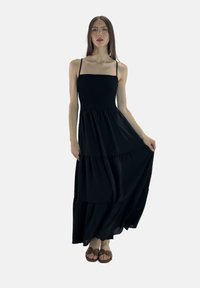 Sort maxikjole med tynde stropper, smockede skørt og lagdelt nederdel. Blødt stof falder elegant. Model med brune sandaler.