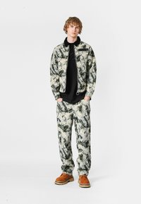 Chaleco y pantalones con un patrón gráfico en tonos verde apagado, crema y negro, combinados con una camiseta negra y zapatos marrones.