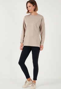 Sweatshirt beige a maniche lunghe con collo rotondo, polsini e orlo a costine, abbinato a jeans skinny neri e sneakers beige chiaro.