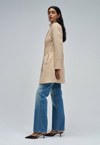 Mujer de perfil con un abrigo largo beige, vaqueros anchos azules y zapatos de tacón bajo de punta negra, sobre un fondo gris liso.